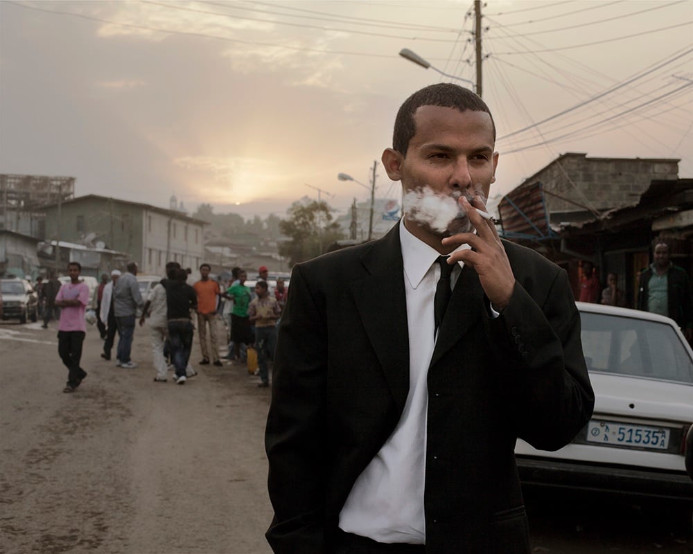 A man in a dark suit on a busy street at dusk, holding a white cloth to his mouth