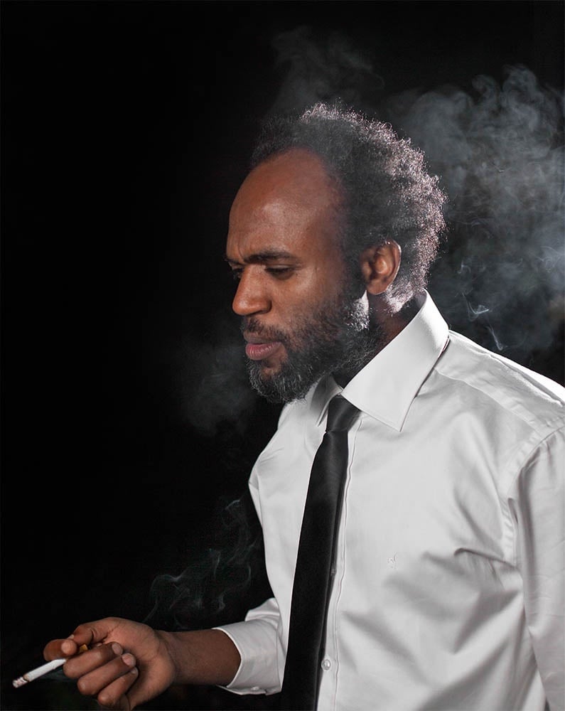 A man in a white shirt and tie smoking a cigarette against a black background
