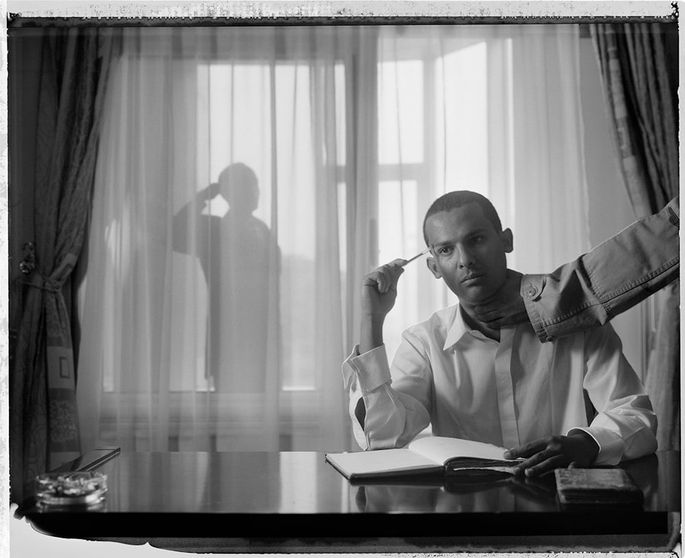 Black and white photograph of a man writing at a desk by a curtained window, a silhouette behind him