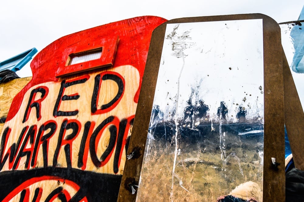 A mirrored shield held up beside a hand-painted Red Warrior banner at the Standing Rock camp