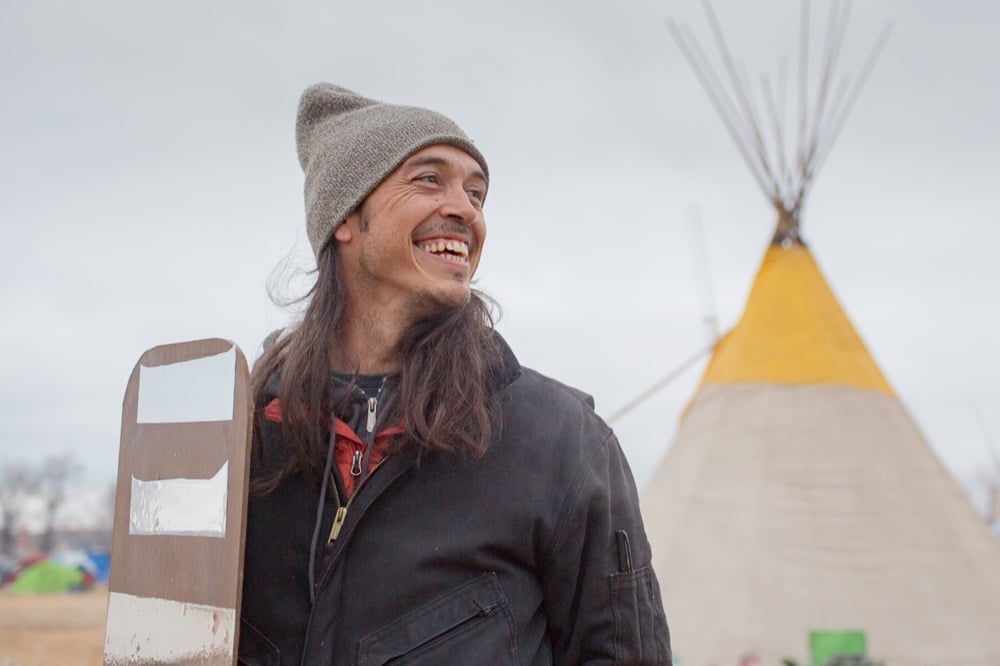Cannupa Hanska Luger smiling in a knit hat beside a mirrored shield, a tipi behind him
